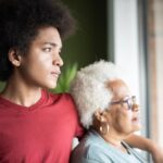 Young person with his arm around an old person, both looking out a window